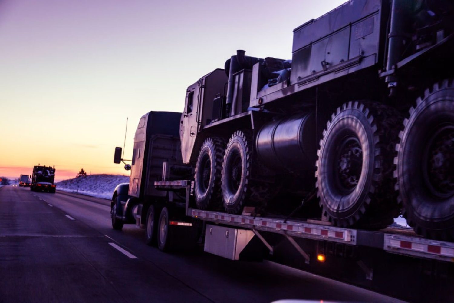 Heading west into a dusky winter sunset, a convoy of semi tractor trailer flatbed trucks loaded with heavy, oversized United States Army tactical armored military land vehicles is hauling the freight along a remote stretch of expressway.